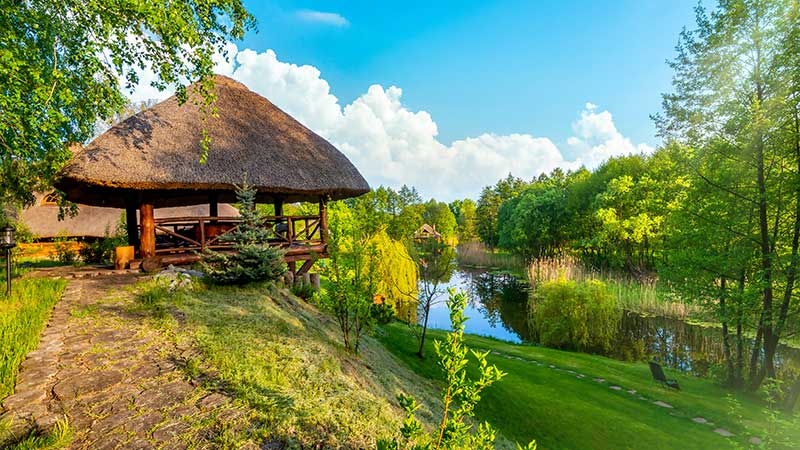 Cabana ecológica com teto de palha ao lado de um lago cercado por árvores e vegetação.