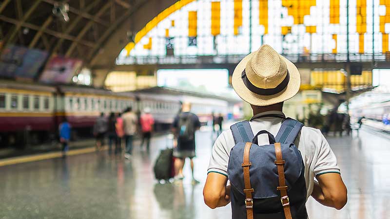 Homem com mochila e chapéu em estação de trem, pronto para viajar de forma sustentável.