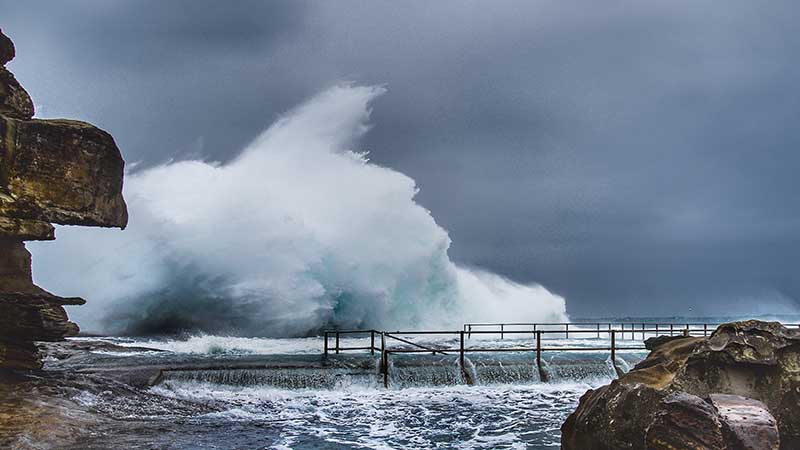 Blog Ambiental • Ondas gigantes atingindo uma área costeira com força, invadindo estruturas de proteção próximas às rochas.