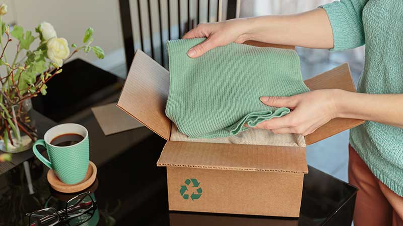 Pessoa embalando roupas em uma caixa de papelão com símbolo de reciclagem, representando a reciclagem de materiais têxteis.