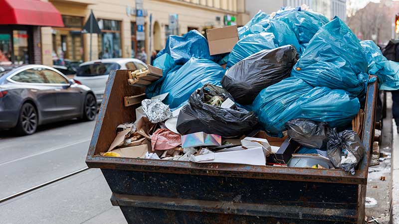 Contêiner de lixo urbano transbordando com sacos plásticos, papelão e resíduos descartados nas ruas.