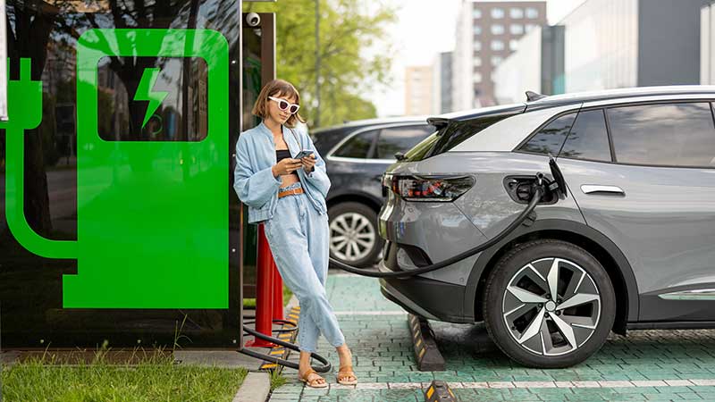 Mulher ao lado de carro elétrico em recarga em estação urbana, simbolizando mobilidade sustentável