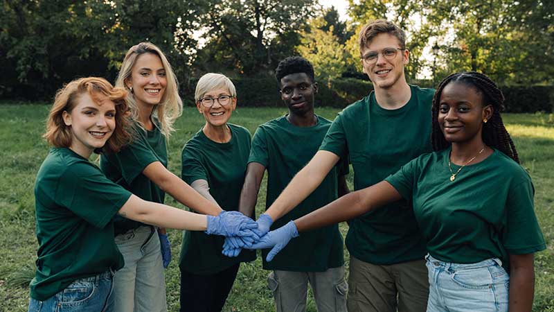 Equipe diversa de voluntários com camisetas verdes unindo as mãos em ação de conscientização ambiental ao ar livre