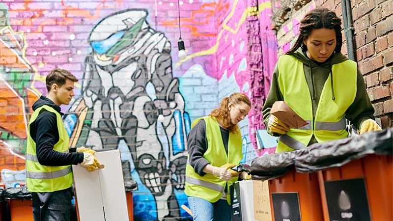 Blog Ambiental - Grupo de jovens com coletes refletivos separando resíduos recicláveis em contêineres, em ambiente urbano com grafite ao fundo