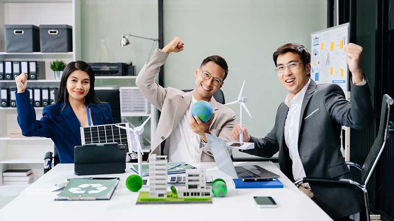 Equipe de profissionais sorrindo e celebrando com maquetes sustentáveis, turbinas e miniaturas do planeta, em ambiente de trabalho ecológico.