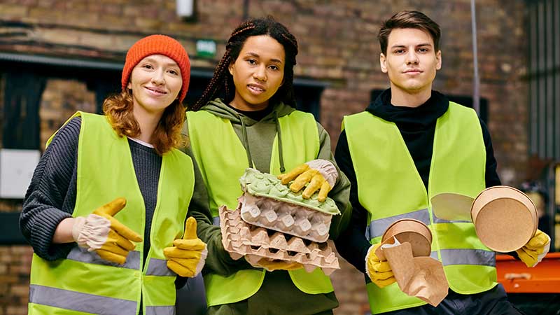Três jovens trabalhadores da reciclagem usando coletes refletivos, sorrindo e segurando embalagens sustentáveis em ambiente de triagem.
