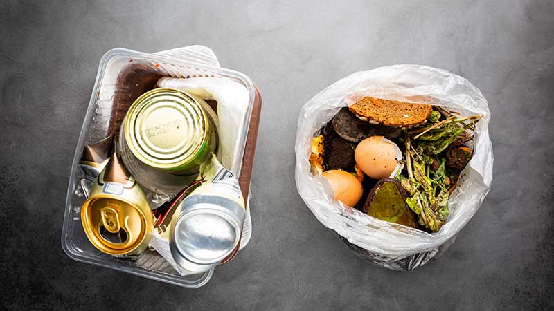 Dois recipientes de lixo contendo alimentos orgânicos e embalagens metálicas descartadas sobre superfície cinza.