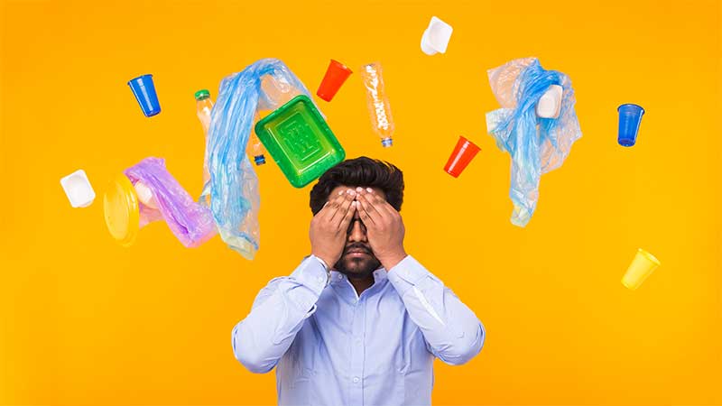 Homem com expressão de estresse cobrindo os olhos, rodeado por embalagens plásticas flutuando sobre fundo amarelo.