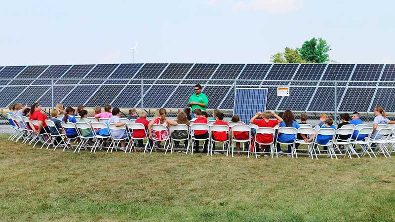 Colaborações para sustentabilidade na educação: professor explica energia solar a crianças diante de painéis fotovoltaicos.