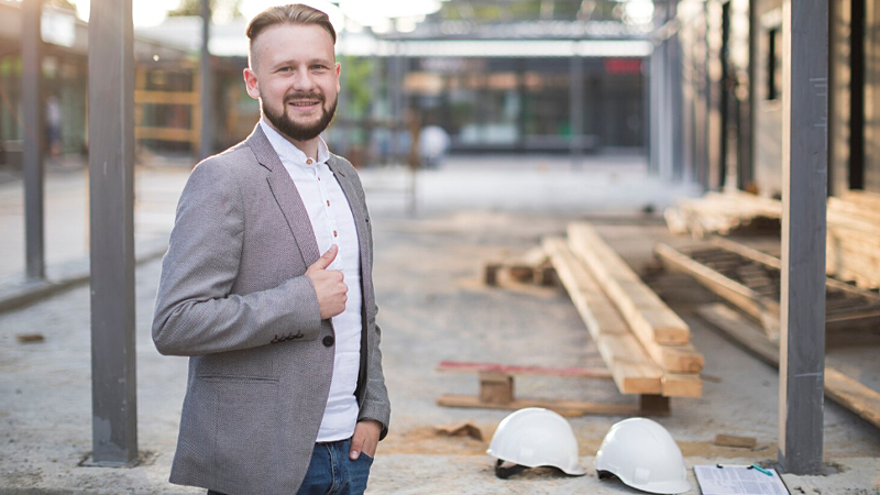 Homem sorridente vestindo blazer claro em um canteiro de obras, simbolizando a liderança e o papel estratégico da gestão na construção de empreendimentos sustentáveis.