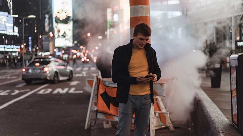 Homem em rua movimentada e poluída à noite, cercado por fumaça, carros e iluminação intensa da cidade.