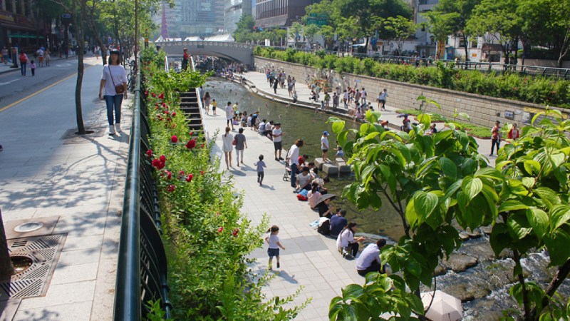 Blog Ambiental • Arroio Cheonggyecheon em Seul, Coreia do Sul, mostrando pessoas caminhando e descansando nas margens revitalizadas após a remoção de uma via expressa elevada, transformada em espaço público verde.