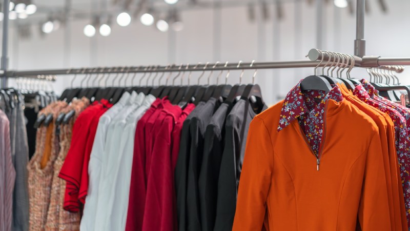 Blog Ambiental • Araras com roupas sofisticadas expostas em loja simbolizam o consumo de tecidos luxuosos impulsionado pela vaidade e pelo desejo de distinção social no capitalismo contemporâneo.